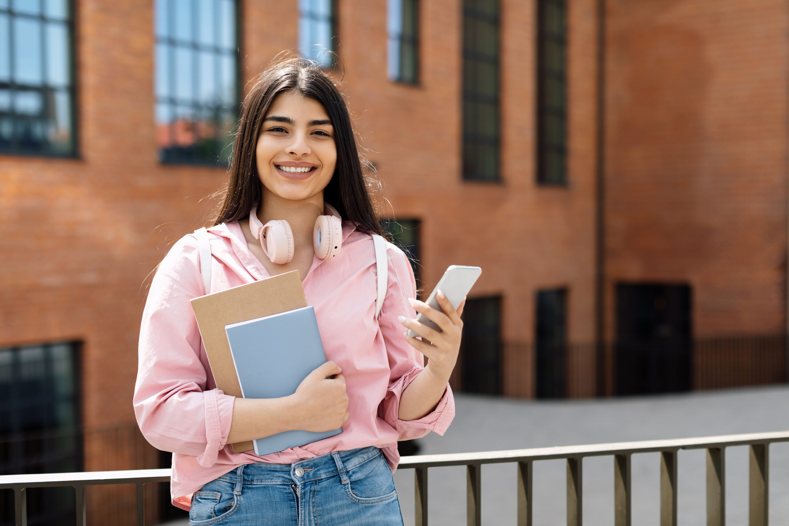 Portrait of cheerful latin student lady with smartphone and workbooks posing outdoors and smiling at camera, female using phone while standing outside near college